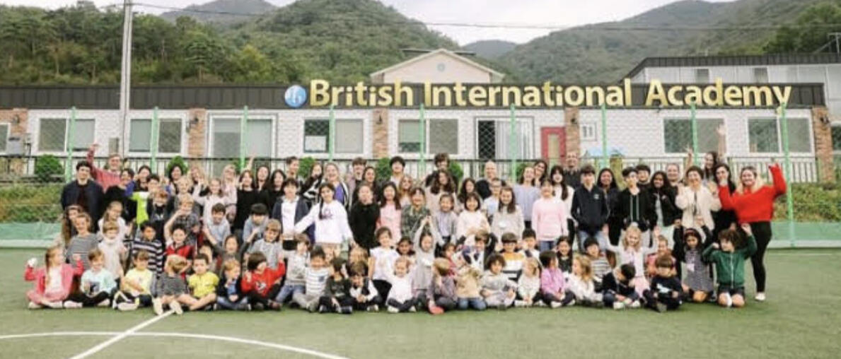Group photo of students, teachers, and staff at British International Academy in Geoje, South Korea, taken on the campus soccer field with the school building and surrounding mountains in the background.